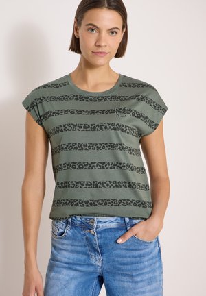 Jeune femme portant un t-shirt vert rayé à imprimé léopard et un jean bleu, une main dans la poche, sur un fond uni.