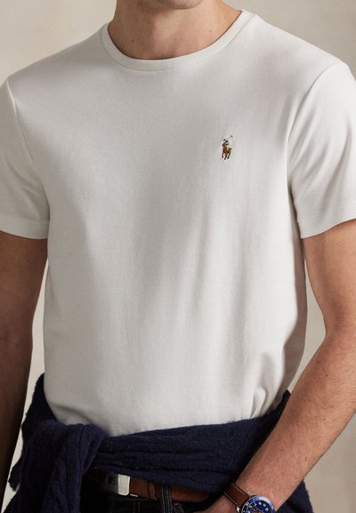 White short-sleeve shirt with polo player logo on chest, navy sweater tied around waist, and wristwatch on left wrist.