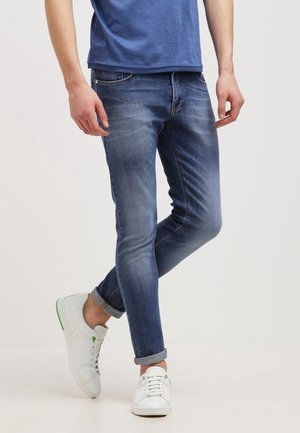 Mężczyzna w podwiniętych niebieskich dżinsach, białych tenisówkach z zielonymi akcentami i niebieskiej koszuli, stojący ze skrzyżowanymi nogami.