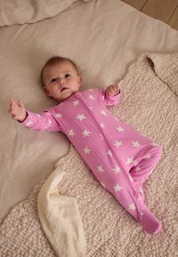 Body rose pour bébé avec des motifs d'étoiles blanches, doté d'une fermeture éclair à l'avant et de manches longues, posé sur une couverture beige texturée.