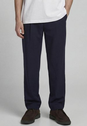 Homme portant un pantalon droit bleu marine foncé, un t-shirt blanc et des chaussures marron foncé, debout avec une main dans la poche.