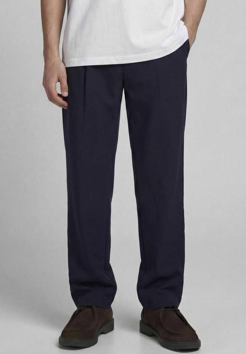 Homme portant un pantalon droit bleu marine foncé, un t-shirt blanc et des chaussures marron foncé, debout avec une main dans la poche.