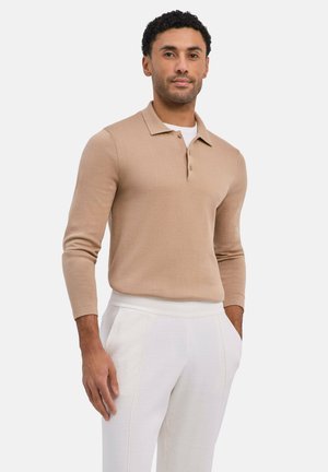 Mann trägt beigefarbenen, langärmeligen Polopullover und weiße Hose, steht mit den Händen in den Taschen vor schlichtem hellem Hintergrund.