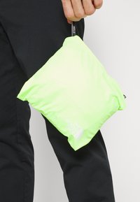 Pouch en verde neón, ligero y packable, con una textura suave, que presenta un logotipo blanco de marca y una pequeña correa negra para llevar.