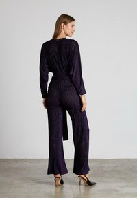 Mono morado texturizado con mangas largas y piernas anchas, que presenta una cintura ceñida. Zapatos de tacón altos negros completan el look.