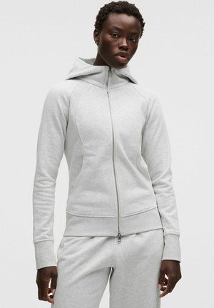 Mujer con sudadera con cremallera gris claro y pantalones deportivos a juego, de pie frente a un fondo liso y claro.