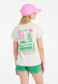 Camiseta beige clara con estampados gráficos coloridos en rosa y verde, con un corte relajado y mangas enrolladas, combinada con shorts verdes.