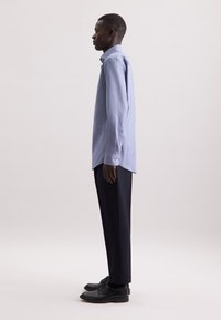 Camicia azzurro chiaro con colletto strutturato, maniche lunghe e bottoni ai polsini visibili, abbinata a pantaloni scuri e scarpe nere.