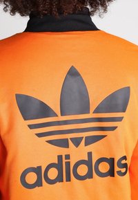 Orange sportjacka med en svart Adidas trefoil-logga och text på baksidan. Materialet verkar slätt, med en strukturerad kragsdesign.