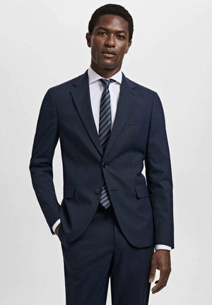 Completo blu navy in tessuto testurizzato, chiusura con un singolo bottone, pantaloni abbinati, indossato con una camicia bianca e una cravatta a righe.