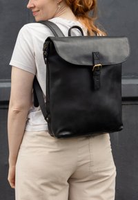 Zaino in pelle nera con chiusura a patta, dotato di una cinghia anteriore e una fibbia, spallacci regolabili e una texture liscia e opaca.