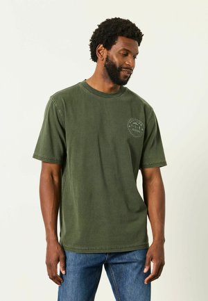 Mann mit kurzen lockigen Haaren und Bart, der ein olivgrünes T-Shirt mit kleinem rundem Logo auf der Brust und blaue Jeans trägt.