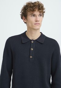 Dunkelblaues, gestricktes Polo-Sweatshirt mit Kragen, das vier braune Knöpfe entlang der Knopfleiste hat. Texturiertes Gewebe, lange Ärmel.