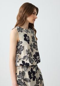 Top senza maniche in tessuto beige con design floreale nero; presenta un colletto rotondo e una vestibilità comoda. Il tessuto testurizzato offre un aspetto naturale.