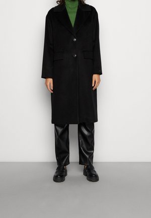 Person wearing a black knee-length coat, green turtleneck, black leather pants, and black chunky loafers, standing against a plain background.