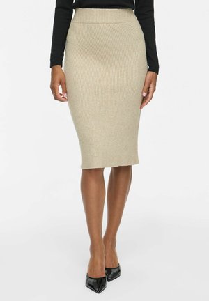 Gonna a tubino beige a coste, indossata con top nero a maniche lunghe e décolleté nere con punta appuntita su una donna in piedi.