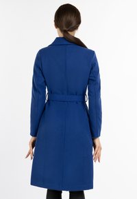Palton albastru din lână cu un design ajustat, mâneci lungi, curea la talie și o croială dreaptă, detalii de coasere vizibile pe spate.