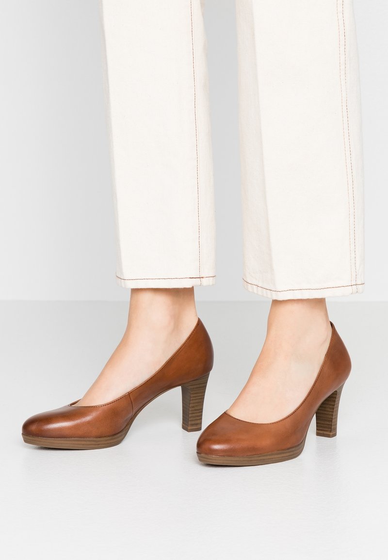 Scarpe alte in pelle marrone con punta arrotondata, dotate di zeppa e tacco impilato texturizzato, abbinate a pantaloni color crema.