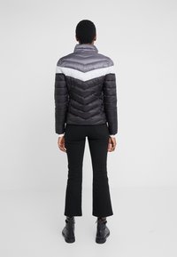 Puffer jacket in black and grey with a white chevron pattern, featuring a high collar and quilted texture. Worn with black flared pants and boots.