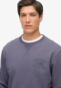 Fioletowa bluza z okrągłym dekoltem, z subtelnym logo "Superdry Athletics" oraz teksturowanym przeszyciem na ramieniu.