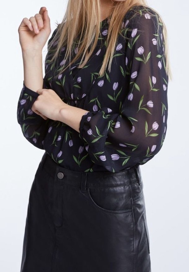 Femme aux longs cheveux blonds portant un chemisier noir transparent à motif tulipe violet et une jupe en cuir noire, ajustant sa manche.