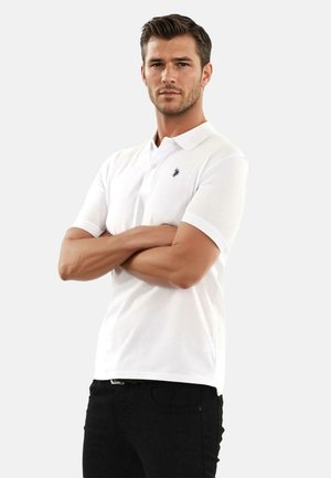 White cotton polo shirt with a folded collar and short sleeves, featuring a small navy logo on the left chest. Worn with black pants.