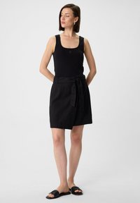 Zwarte mouwloze top, zwarte wikkelrok met een strikceintuur en zwarte instapsandalen, met gladde texturen en een eenvoudig, minimalistisch ontwerp.