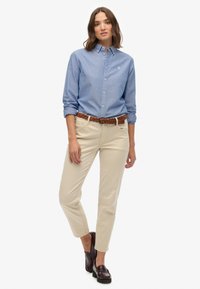 Kobieta stoi w jasnoniebieskiej koszuli zapinanej na guziki, beżowych spodniach z brązowym plecionym paskiem i ciemnobrązowych półbutach na białym tle.