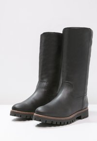 Paire de grandes bottes en cuir noir avec semelles épaisses à crampons et garniture marron, debout sur une surface blanche contre un fond uni.