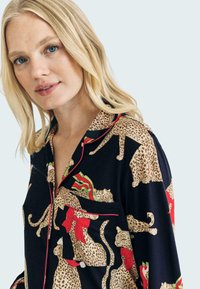 Black pajama top featuring a leopard print design, festive red bows, and green accents. Includes a pocket with a matching pattern and red piping.