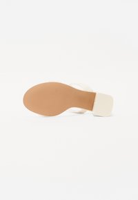 Sandal de tacón alto beige con tacón bloque, suela texturizada color bronce, y tiras cruzadas. Acabado suave con un diseño minimalista.