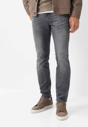 Mand iført grå slim fit jeans, brune ruskindssneakers med hvide såler, lysebrun jakke og beige sweater, stående på hvid baggrund.