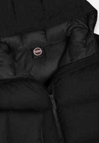 Melns pūkains jakas ar rāvējslēdzēju un kapuci. Iezīmējas ar quilted daļām un Colmar Originals etiķeti iekšējā apkaklē.