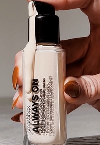 Fond de teint Smashbox Always On dans une bouteille en verre transparent avec une pompe noire, délivrant un liquide beige clair. Présente un texte noir en gras sur l'étiquette.