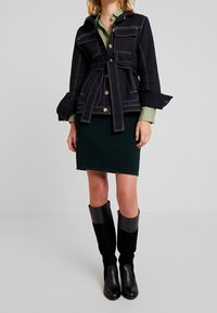 Svart denimjacka med guldknappar, bälte och fyra framfickor. Kombineras med en ribbad grön kjol och svarta knähöga stövlar.