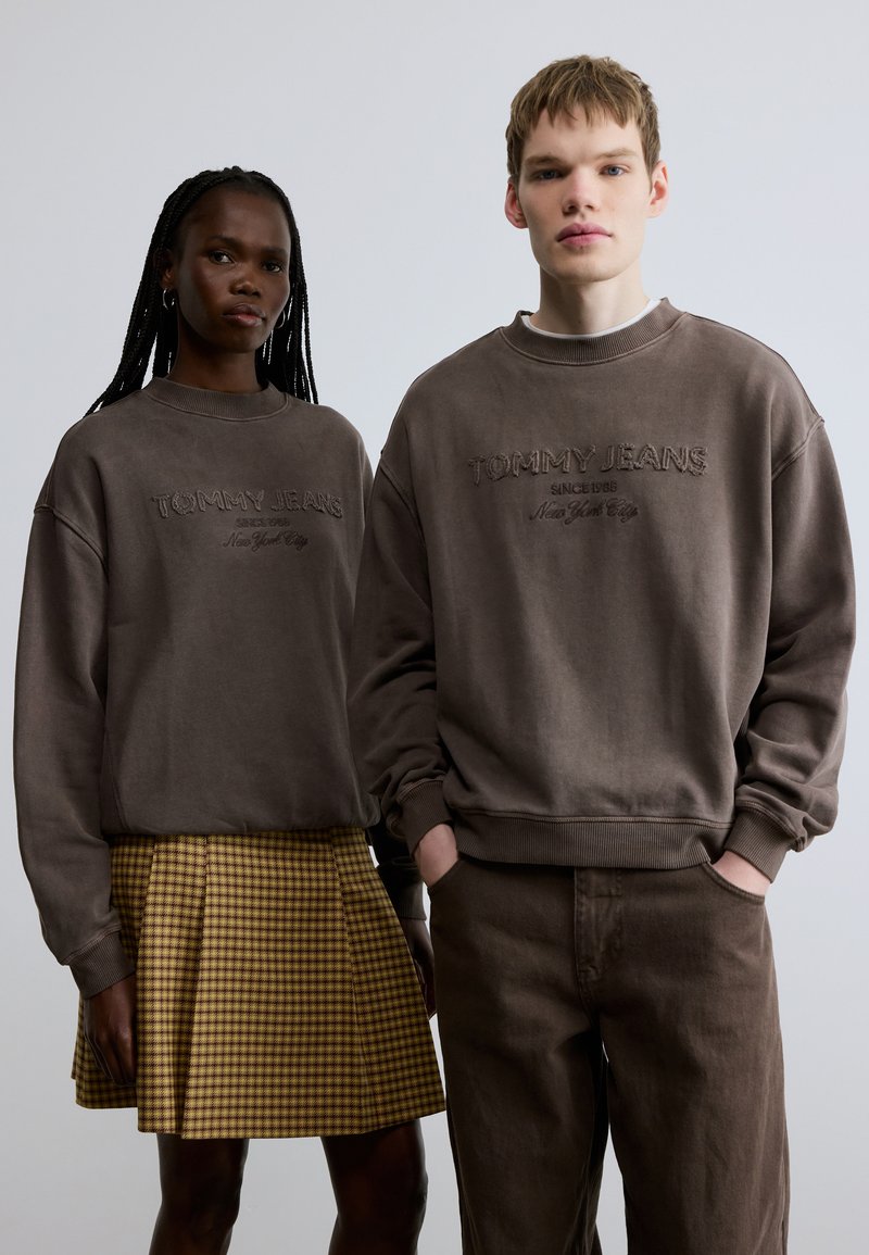 Sudaderas marrones de gran tamaño con el logo bordado "TOMMY JEANS"; la modelo lleva una falda plisada de cuadros amarillos. La textura parece suave y casual.