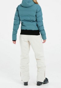 Tiefblaue Pufferjacke mit Kapuze, gerippter schwarzer Saum und Bündchen, kombiniert mit cremefarbenen Schneehosen mit Seiteneingriffen und schwarzen Skistiefeln.