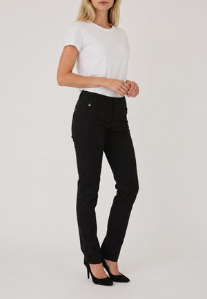 Femme portant une chemise blanche à manches courtes, un pantalon noir slim et des chaussures noires à talons hauts, debout devant un fond uni.