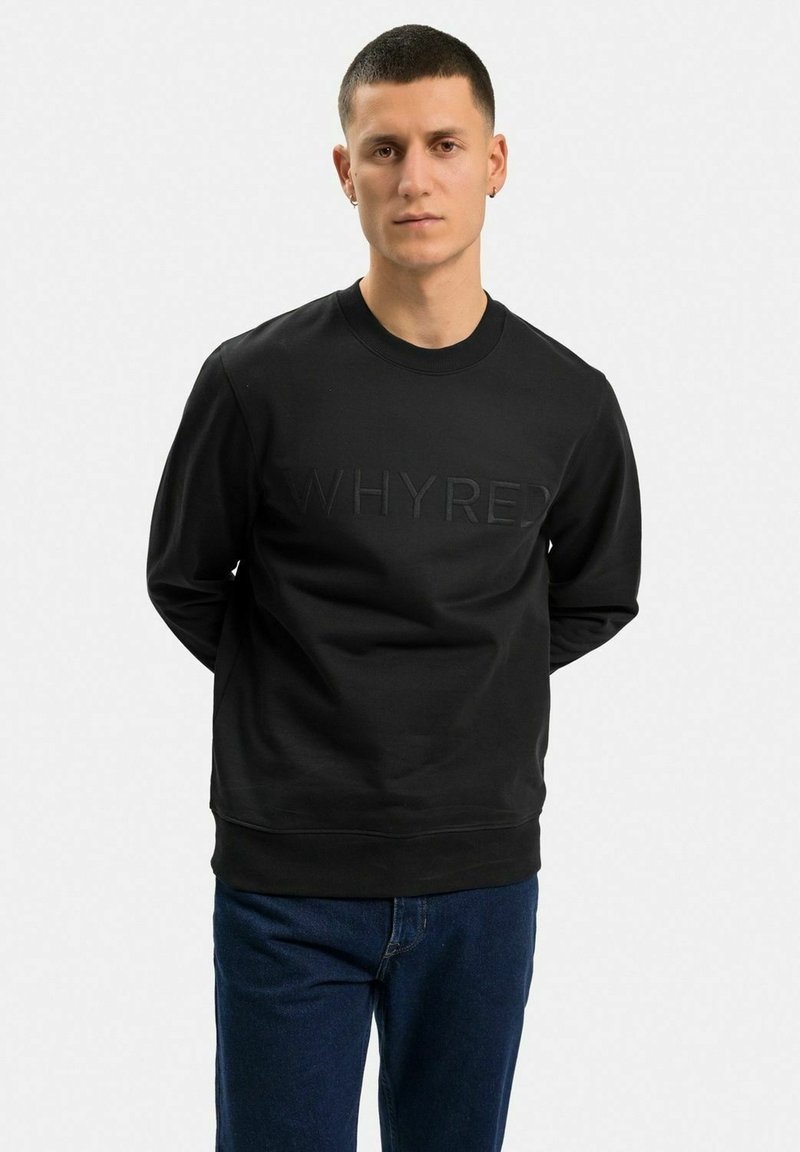 Svart sweatshirts med präglad "WHYRED" text framtill, rund halsringning, långa ärmar och ribbade manschetter, matchat med blå jeans.