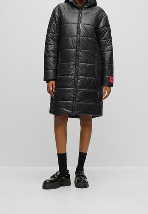 Person trägt eine knielange schwarze gesteppte Steppjacke mit Kapuze, vorderem Reißverschluss, Seitentaschen, schwarzen Socken und klobigen schwarzen Loafern.