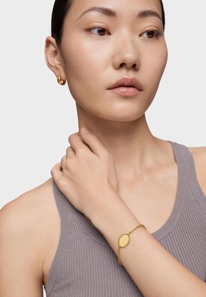 Pulsera de oro con un charm ovalado en una delicada cadena. El modelo lleva puesta una camiseta gris de canalé y pendientes de aro dorados, con la mano descansando sobre su cuello.