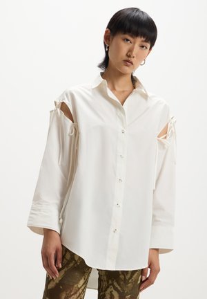 Camicia bianca con spalle scoperte, dettagli annodati, maniche lunghe, texture liscia e vestibilità rilassata. Abbinata a pantaloni verdi a fantasia.
