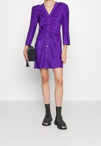 Lila knappad klänning med långa ärmar, rynkade detaljer och ett strukturerat mönster. Matchas med svarta ankle boots och en liten handväska.