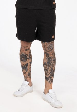 Nogi z czarnymi tatuażami kwiatowymi i symbolami, w czarnych szortach i białych tenisówkach, stojące na białym tle.