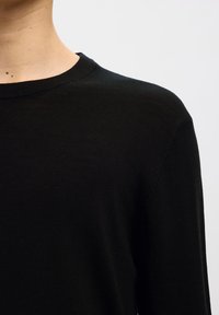 Közeli kép egy személyről, aki egyszínű fekete, kerek nyakú pulóvert visel, megmutatva a nyakat, vállat és a mellkas felső részét világos háttér előtt.