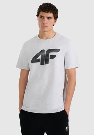 Młody mężczyzna z krótkimi kręconymi włosami, ubrany w jasnoszarą koszulkę z dużym czarnym logo "4F" oraz czarne spodnie z logo "4F" na lewej nogawce.