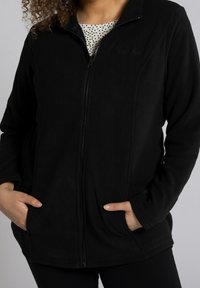 Giacca in fleece nera con zip e tasche laterali, caratterizzata da una texture morbida e un logo sul petto. Progettata per offrire calore e comfort.