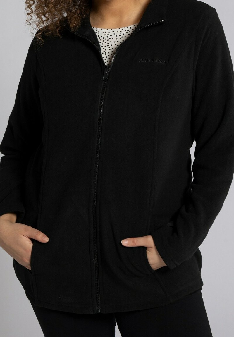 Giacca in fleece nera con zip e tasche laterali, caratterizzata da una texture morbida e un logo sul petto. Progettata per offrire calore e comfort.