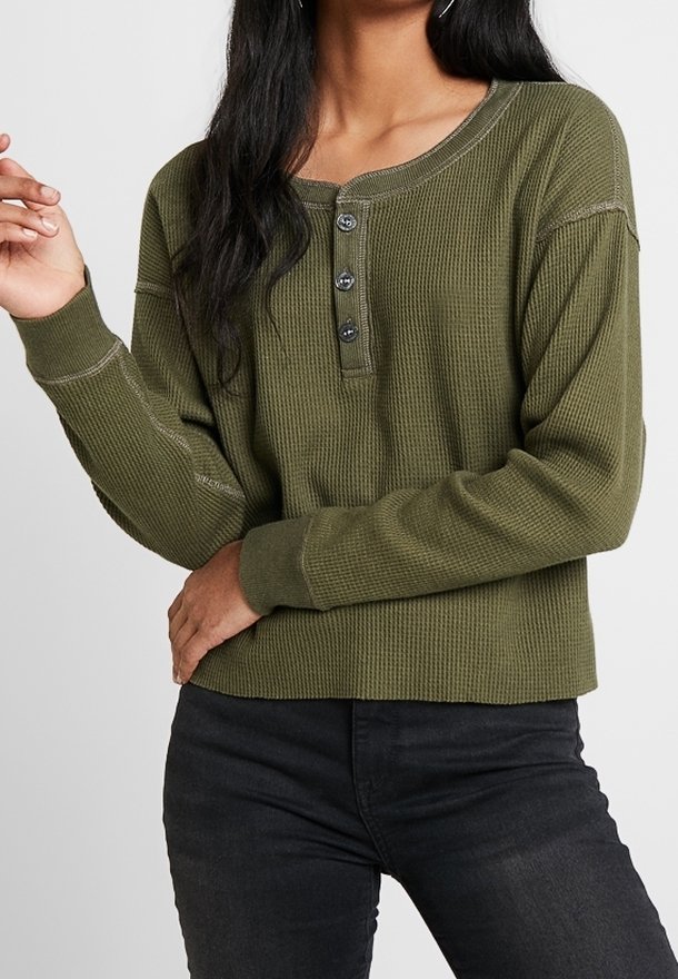 Femme portant un t-shirt henley en tricot gaufré olive vert à manches longues avec trois boutons et un jean noir, posant avec une main levée et l'autre sur sa taille.