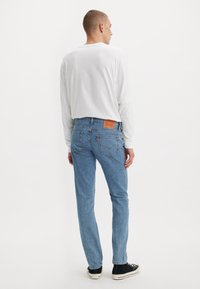 Man staat met zijn rug naar de camera, gekleed in een wit overhemd met lange mouwen, lichtblauwe jeans en zwarte sneakers, tegen een witte achtergrond.